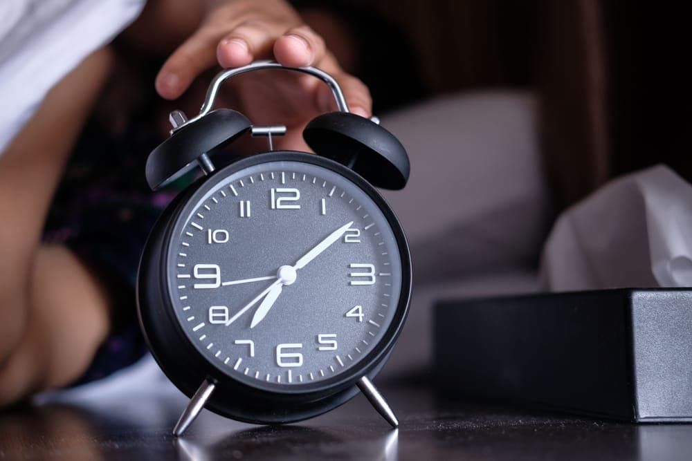 A hand reaching for an analog alarm clock on a nightstand beside a bed