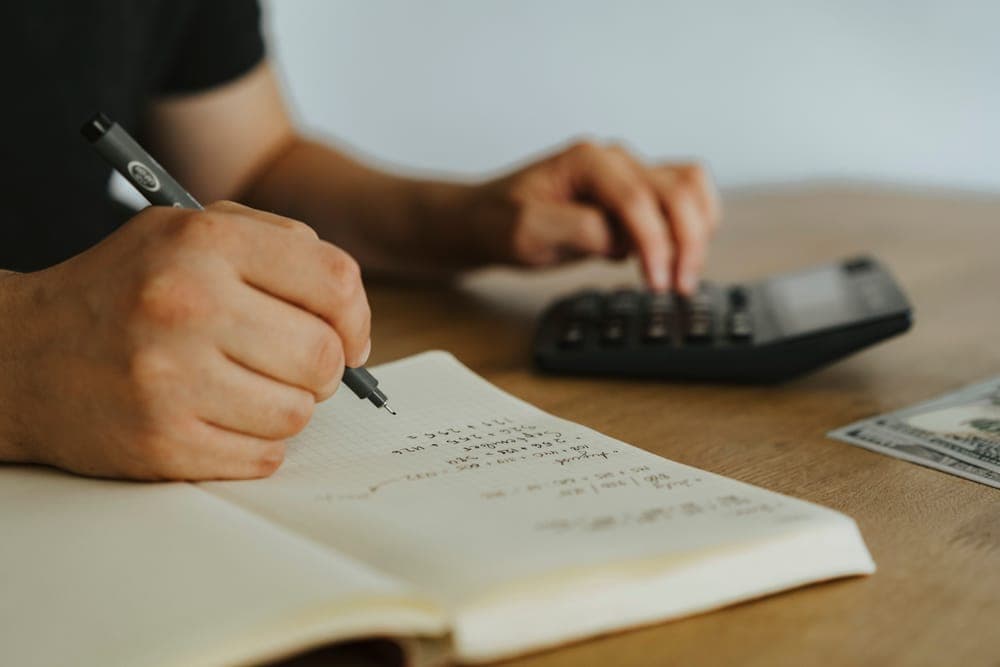 A hand writing figures in a notebook beside a calculator on a wooden desk