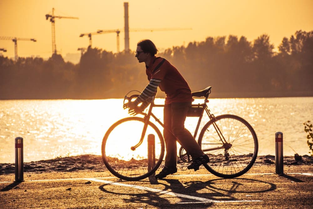 A man standing astride a road bike on a riverside path at golden hour