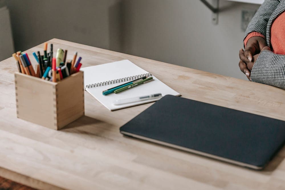 A wooden desk with a notebook, pens and a closed laptop