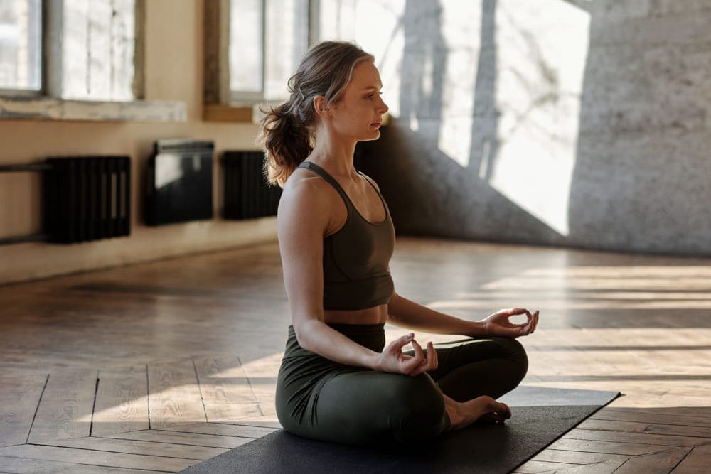 A woman sitting cross-legged in meditation on a sunlit wooden floor