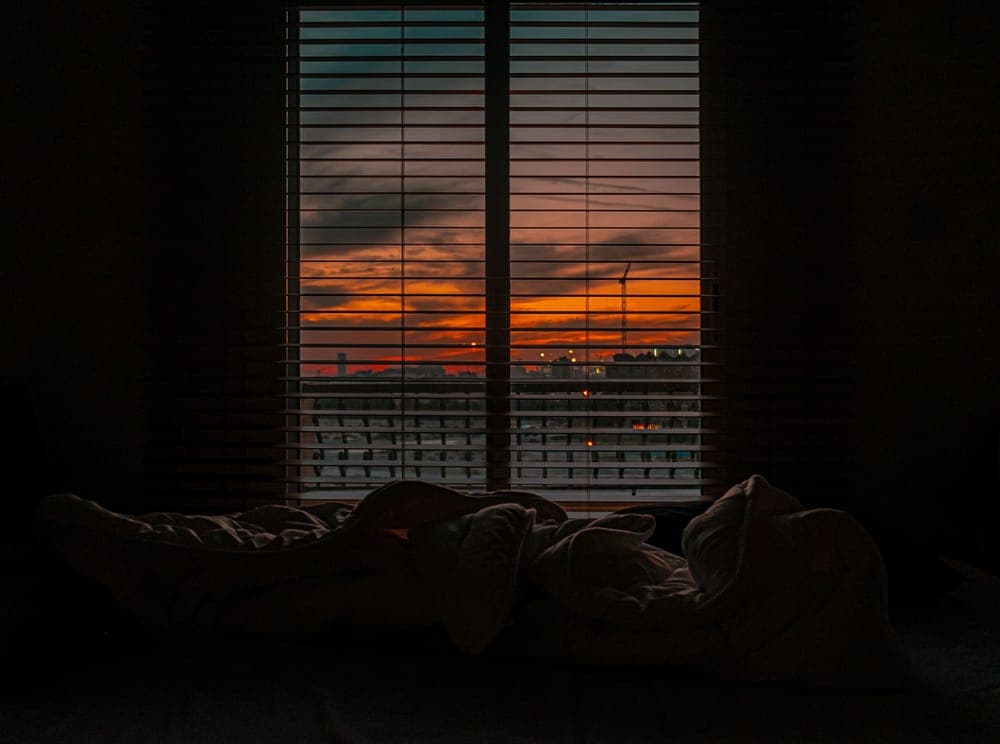 A dark bedroom with a bed in front of a window at dusk