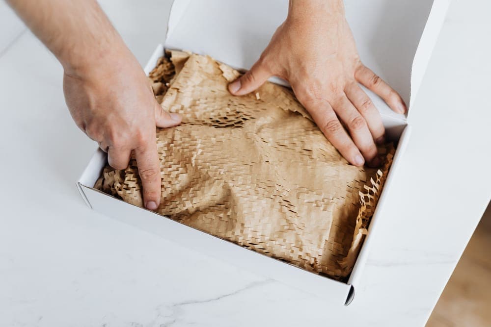Hands packing kraft paper into an open white box on marble