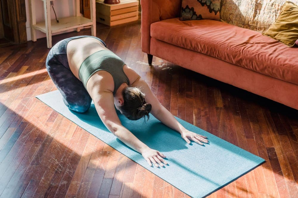 A woman in child's pose on a mat on a sunlit wooden floor