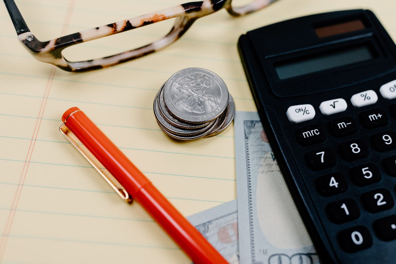 A calculator, a stack of quarters, a pen and glasses on a yellow legal pad