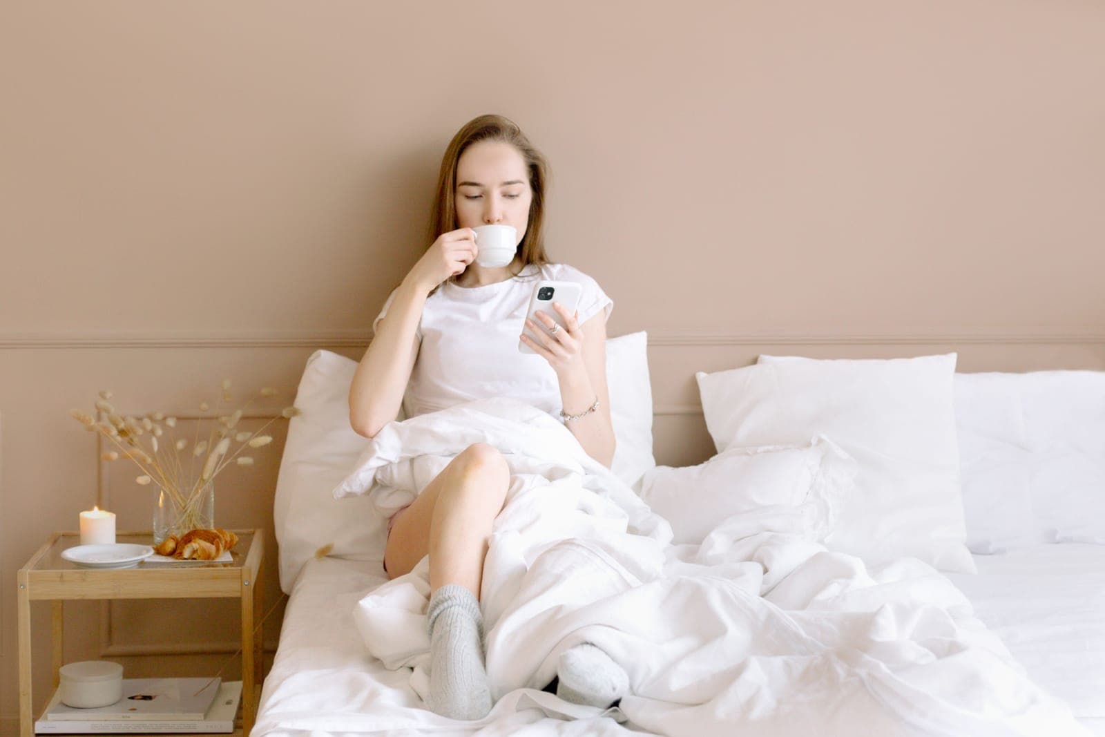 A woman sitting up in bed with a cup, looking at her phone beside a wooden side table
